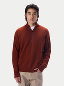 A man wearing a rust-brown quarter-zip knit sweater over a white undershirt and beige trousers, standing against a light gray studio background.