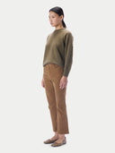 A model is shown in profile wearing a muted olive green knit sweater with a textured cable pattern on one sleeve, paired with brown trousers and flat shoes, against a light gray background.