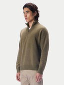 A man wearing a muted olive green quarter-zip knit sweater and beige trousers, photographed against a clean light gray studio background.