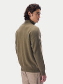 A man wearing a muted olive green knit turtleneck sweater, shown from a side-back angle against a clean light gray studio background.