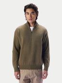 A man wearing a muted olive-brown quarter-zip knit sweater over a white undershirt and beige trousers, standing against a plain light gray studio background.