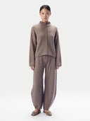A model stands against a light gray studio background wearing a matching taupe knit two-piece set consisting of a mock-neck long-sleeve top with a central braided detail and wide-leg trousers.