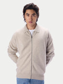 A man wearing a light beige zip-up knit cardigan over a white t-shirt and blue jeans, photographed against a solid light gray background.