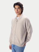 A man wearing a light beige zip-up knit sweater over a white t-shirt and blue jeans, photographed against a solid light gray background.