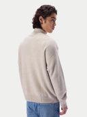 A man wearing a light beige knit mock neck sweater, shown from a three-quarter back view against a plain light gray studio background.