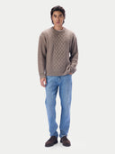A man modeling a taupe cable-knit crewneck sweater, paired with light blue jeans and dark brown suede loafers, against a solid light gray background.
