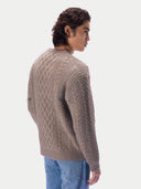 A man wearing a taupe cable-knit sweater and blue jeans, shown from a side-back angle against a light gray studio background.