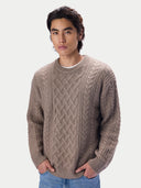A man wearing a taupe cable-knit crewneck sweater over a white undershirt and blue jeans, standing against a light gray studio background.