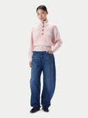 A model stands against a light gray studio background wearing a pale pink mock-neck knit sweater with brown buttons, loose-fit blue jeans, and dark loafers.