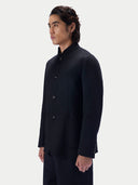 A man wearing a dark navy or black mandarin-collar jacket with front buttons, shown in a side-profile studio shot against a light gray background.