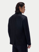 A man shown from the back wearing a black high-neck jacket and matching trousers against a light gray studio background.