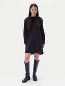 A model wearing a black long-sleeve knit dress with an asymmetrical draped tie detail at the neckline, paired with dark knee-high boots, against a plain light gray studio background.