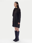 A model is shown in profile wearing a black knit wrap dress with a draped shawl collar, paired with black knee-high boots, against a plain light gray studio background.