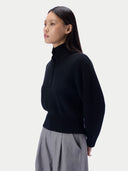 A woman in a side profile wearing a black ribbed mock-neck quarter-zip sweater and gray pleated trousers, set against a solid light gray background.