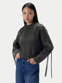 A woman modeling a dark olive green ribbed knit sweater with unique textured shoulder details and long braided tie details on the sleeves, paired with blue jeans against a light gray background.