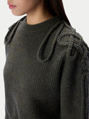 Close-up of a person wearing a dark olive green ribbed knit sweater with decorative knotted detailing on the shoulders, against a plain light background.