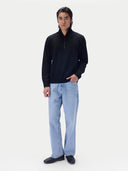 A man modeling a black long-sleeve quarter-zip pullover sweater paired with light blue jeans and black loafers, standing against a solid off-white studio background.