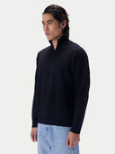 A man wearing a black quarter-zip knit sweater and light blue jeans, photographed against a clean light gray studio background.