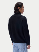 A man shown from the back wearing a black mock-neck knit sweater and light blue jeans against a plain light gray studio background.