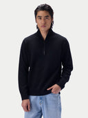 A man wearing a black quarter-zip mock neck sweater and light blue jeans, standing against a light gray studio background.