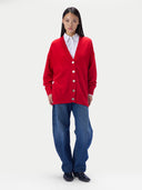 A model stands against a light gray studio background wearing a bright red button-front cardigan over a white collared shirt, with loose blue jeans and black shoes.