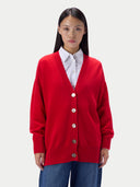 A model wearing a vibrant red knit cardigan over a white collared shirt and blue jeans, photographed against a solid light gray background.