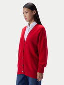 A woman wearing a bright red knit cardigan over a white collared shirt and blue jeans, photographed against a clean light gray studio background.