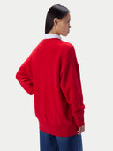 A woman shown from the back wearing a bright red knit sweater over a white collared shirt and blue jeans, photographed against a plain light gray studio background.