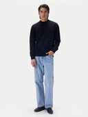 A man modeling a black long-sleeve mock neck sweater paired with light blue jeans and dark loafers, standing against a solid off-white background.