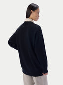 A woman shown from the back wearing a dark navy knit sweater over a cream collared shirt and light beige trousers, photographed against a clean light gray studio background.