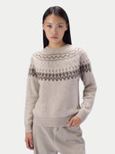 A woman modeling a light beige knit sweater with a dark brown Fair Isle pattern across the chest and shoulders, paired with light beige trousers, against a solid light gray background.