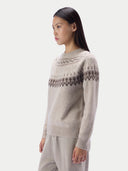 A woman wearing a light beige knit sweater with a dark gray geometric Fair Isle pattern across the chest and shoulders, paired with matching light beige trousers, photographed against a solid light gray background.