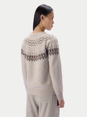 A woman shown from the back wearing a cream-colored knit sweater with a dark gray geometric Fair Isle pattern across the shoulders and upper back, paired with light gray trousers, against a solid off-white background.
