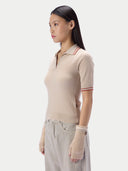 A woman shown in side profile wearing a beige ribbed short-sleeve polo-style knit top with thin red and white striped trim at the collar and sleeve cuffs, paired with light beige knit fingerless gloves and light gray pants, against a plain light gray studio background.
