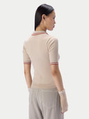 A woman shown from the back wearing a beige ribbed short-sleeve knit top with thin red and white striped trim at the collar and sleeve cuffs, paired with light gray trousers against a plain light gray studio background.
