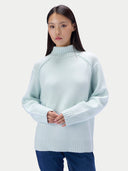 A woman wearing a pale mint green mock-neck knit sweater and dark blue jeans, standing against a solid light gray background.