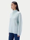 A woman wearing a pale icy blue mock neck knit sweater and dark blue jeans, shown in a side profile against a light gray studio background.