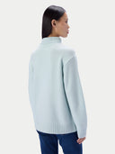 A woman shown from the back wearing a pale icy blue mock-neck knit sweater and dark blue jeans against a light gray studio background.