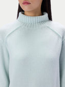A close-up of a model wearing a pale mint green ribbed mock neck sweater against a light off-white background.