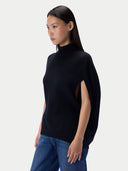 A woman wearing a black sleeveless mock-neck knit top with side cutouts and blue jeans, photographed against a light gray studio background.