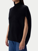 A woman wearing a black sleeveless mock-neck knit sweater with side slits, paired with blue jeans, shown against a light gray studio background.