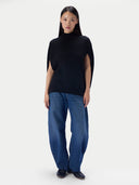 A model stands against a light gray studio background wearing a black mock-neck knit top with dramatic shoulder cutouts, wide-leg blue jeans, and dark loafers.