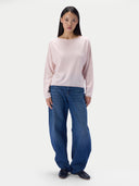 A model stands against a plain light gray studio background wearing a pale pink long-sleeve knit top, loose blue jeans, and dark shoes.
