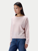 A woman modeling a light pink knit sweater with a wide boat neckline, paired with blue jeans, against a plain light gray studio background.