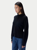 A woman wearing a black long-sleeve knit cardigan with a buttoned front and blue jeans, photographed against a plain light gray studio background.