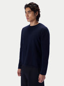 A man shown in three-quarter profile wearing a navy blue crewneck knit sweater with a rolled neckline and dark trousers, against a light gray studio background.