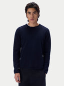 A man wearing a navy blue long-sleeve crewneck knit sweater over a white t-shirt and dark trousers, standing against a light gray studio background.