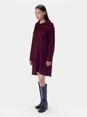 A model wearing a long-sleeved, knee-length draped dress in deep burgundy, paired with black knee-high boots, against a plain light gray studio background.
