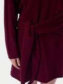 A close-up of a long-sleeve wrap dress or robe in deep burgundy, featuring a self-tie belt and a soft draped neckline, shown on a model against a light gray background.