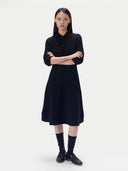 A model stands against a light gray studio background wearing a black three-quarter sleeve polo-knit midi dress, paired with black socks and black leather shoes.
